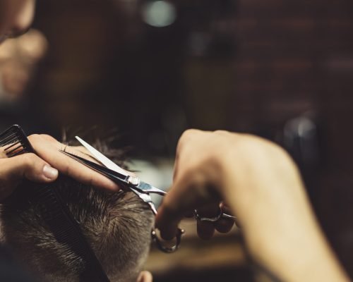 Anonymous stylish barber with tattoos cutting hair of male client in chair.