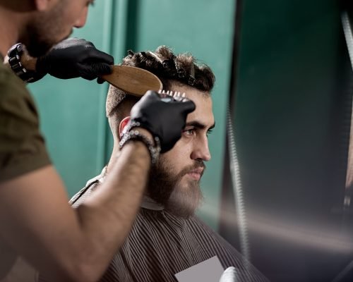 Handsome young man with beard sits at a barber shop. Barber in black gloves shaves hairs at the side. .
